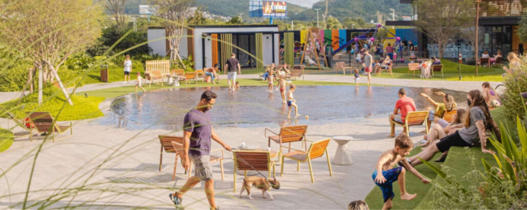 O Passeio Sapiens, novo empreendimento no norte da Ilha de Santa Catarina, será um centro de compras e de convivência a céu aberto. Na foto, o térreo do Passeio Primavera. Ao centro, um lago e um homem passeando com seu cão. Ao lado direito, crianças brincando em meio à grama verde.