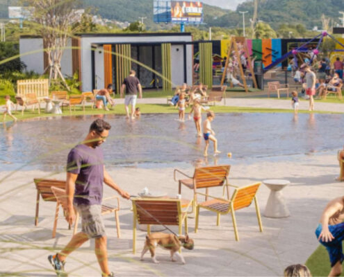 O Passeio Sapiens, novo empreendimento no norte da Ilha de Santa Catarina, será um centro de compras e de convivência a céu aberto. Na foto, o térreo do Passeio Primavera. Ao centro, um lago e um homem passeando com seu cão. Ao lado direito, crianças brincando em meio à grama verde.